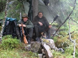 Dag Kjelsaas og Knut Brevik sitter med hvert sitt våpen ved en granlegg. De fyrer bål og holder hvert sitt våpen.